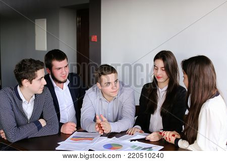 Young and active male and female administrators of company together read annual report and laugh with each other's smiles. Friendly company of professionals solves important issues and problems together, sitting at table in office of company during workin