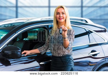 Young happy smiling blonde business woman driver holding auto keys in front of a luxury car