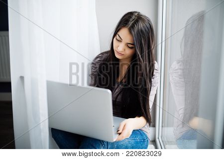 Beautiful Young Woman Sitting On Windowsill And Using Laptop