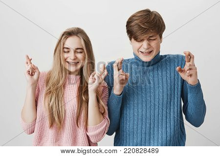 Young caucasian couple with closed eyes, frowning their faces and crossing fingers wishing something posing against gray background. Emotional girl and boy hope for good. Body language