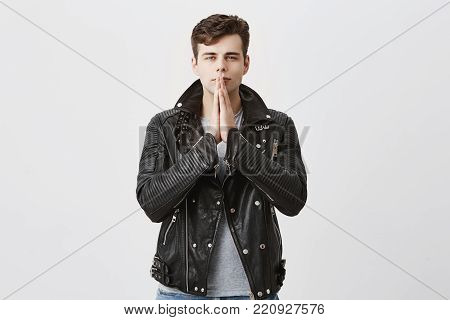Serious concentrated handsome man in black leather jacket keeps palms together, prays for well being of his family, hopes for better, has strong belief. Trendy good-looking male student worries about his future, asks for good luck