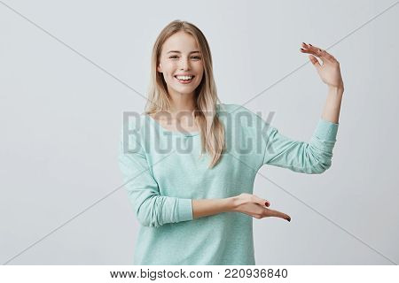 Waist-up portrait of cheerful lovely caucasian female model with blonde hair smiling broadly with white teeth demonstrating size of the box with present, gesturing actively. Body language and face expression