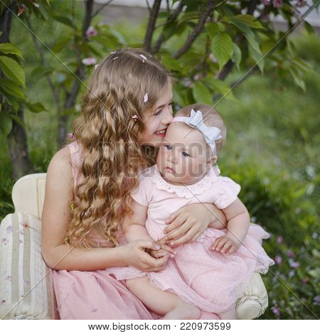 Two happy little sisters love and hug in beautiful park. Cute little girls in pink dress play in spring garden. Adorable child walk and laugh on green meadow