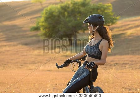 Girl On A Mountain Bike On Offroad, Beautiful Portrait Of A Cyclist At Sunset, Fitness Girl Rides A 