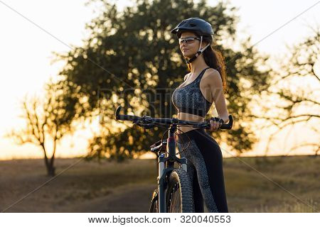 Girl On A Mountain Bike On Offroad, Beautiful Portrait Of A Cyclist At Sunset, Fitness Girl Rides A 