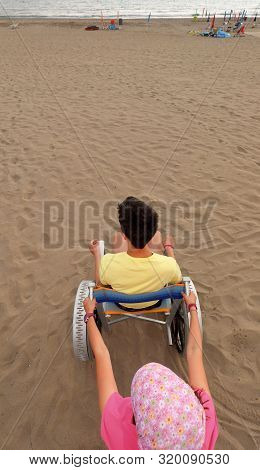 Little Girl Pushes The Wheelchair On The Sandy Beach With A Young Boy In Summer