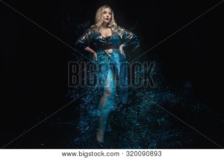 Full length plus size model. Mixed blue lights effect. Young woman in shiny evening dress with sequins. The posing with hands on the hips. Black background. Fashion shot.