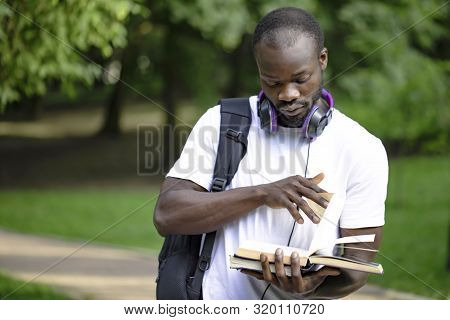Portrait Of Black Student Youngster With Daypack Flipping Book Pages In Campus Park