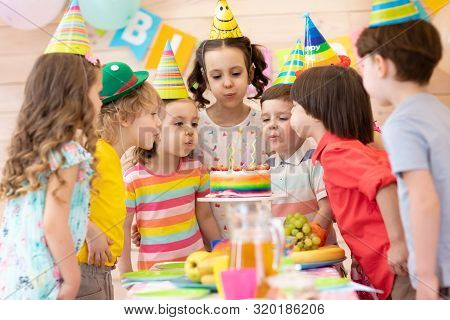 Group Of Adorable Kids 3-5 Years Gathered Around Festival Table. Children Blow Out Candles On Cake. 