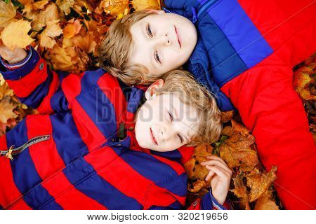 Two Little Kid Boys Lying In Autumn Leaves In Colorful Fashion Fall Clothing. Happy Healthy Siblings