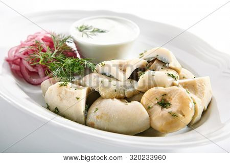 Pickles set with homemade pickled milk mushrooms, marinated red onion rings and sour cream on white restaurant plate isolated. Salted Lactarius resimus mushroom with oil and fresh chopped dill