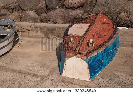 Small Colorful Wooden Fishing Boat On A Streetwalk Of The Port. Up Side Down, Visible Wheels For Tra