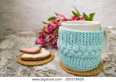 White Cup Of Tea In A Knitted Blue Case With Pink Flowers Tulips And Sweets, With Gingerbread On A T