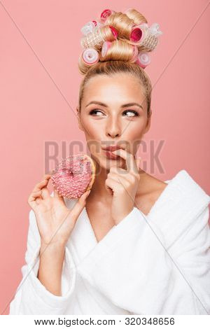 Portrait of caucasian young housewife in housecoat with hair curlers smiling while eating sweet donut isolated over pink background