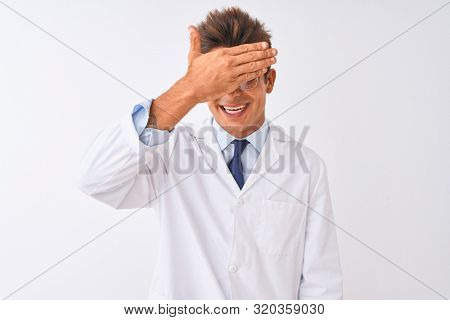 Young handsome sciencist man wearing glasses and coat over isolated white background smiling and laughing with hand on face covering eyes for surprise. Blind concept.