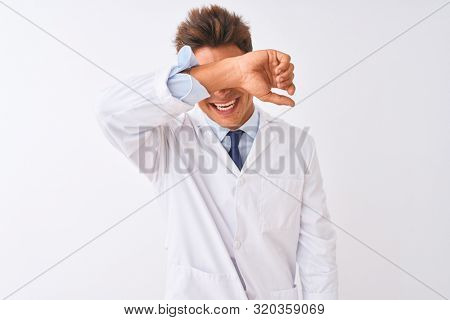 Young handsome sciencist man wearing glasses and coat over isolated white background covering eyes with arm smiling cheerful and funny. Blind concept.