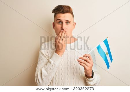 Young handsome man holding Argentinian Argentina flag over isolated white background cover mouth with hand shocked with shame for mistake, expression of fear, scared in silence, secret concept