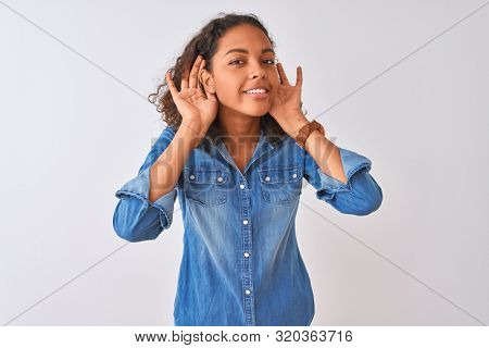 Young brazilian woman wearing denim shirt standing over isolated white background Trying to hear both hands on ear gesture, curious for gossip. Hearing problem, deaf