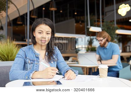 Portrait Of Young Woman In Casual Clothing Looking At Camera While Sitting At The Table And Writing 