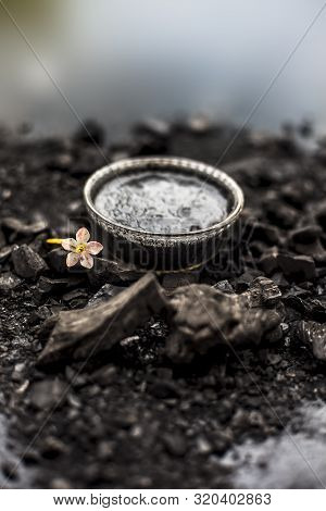 Close Up Of Activated Charcoal In A Glass Bowl On The Wooden Surface Along With Some Raw Powder Of C
