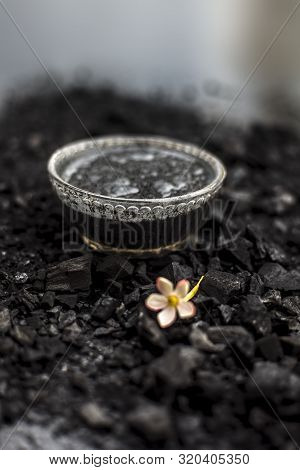 Close Up Of Activated Charcoal In A Glass Bowl On The Wooden Surface Along With Some Raw Powder Of C