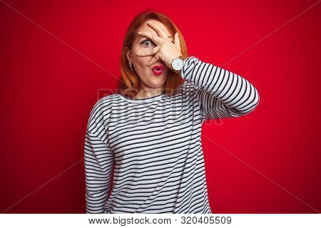 Young redhead woman wearing strapes navy shirt standing over red isolated background peeking in shock covering face and eyes with hand, looking through fingers with embarrassed expression.