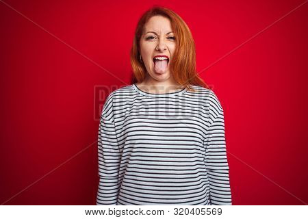 Young redhead woman wearing strapes navy shirt standing over red isolated background sticking tongue out happy with funny expression. Emotion concept.