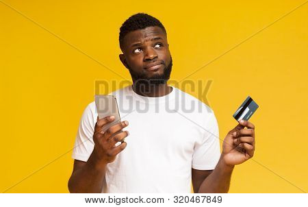 Do I Longer Need A Credit Card. Hesitant African American Man Holding Smartphone And Card, Looking U