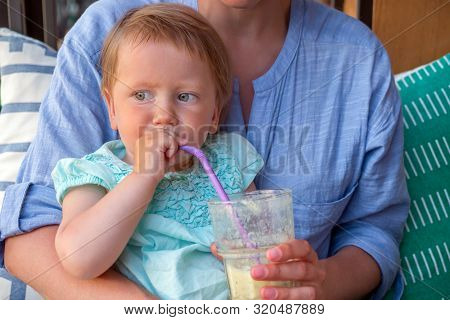 Blond Baby Girl With Blue Eyes Sits On Mother Lap. Little Girl Tastying Detox, Fresh Juice, Vegetalb