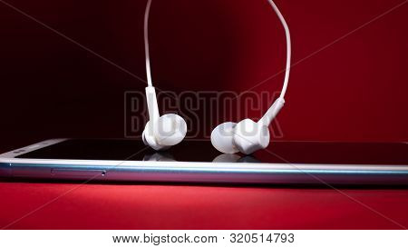 Concept: Listen To Music From The Phone.white Headphones Hanging Over A Smartphone On A Red Backgrou
