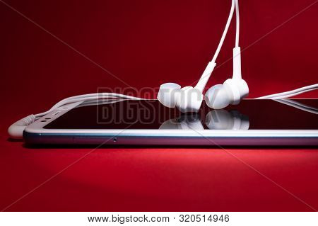Concept: Listen To Music From The Phone.smar Tphone With White Headphones On A Red Background Closeu