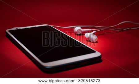 Concept: Listen To Music From The Phone. Smartphone With Headphones In The Rays Of Light On A Red Ba