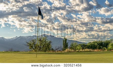 Panorama Focus On The Flagstick Of A Golf Course Against Trees Mountain And Cloudy Sky