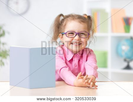 Happy kid girl in eyeglasses reading with standing book on table