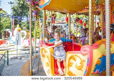 Little Blonde Girl On Old French Carousel In A Holiday Park. Charming Cute Little Girl In A Dress Is