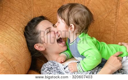 Baby Bites Moms Nose. Happy Mother With Baby. A Boy Bites His Mothers Nose On The Couch. Having Fun 