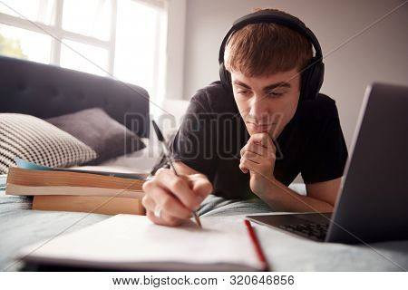 Male College Student Wearing Headphones Lies On Bed In Shared House Working On Laptop