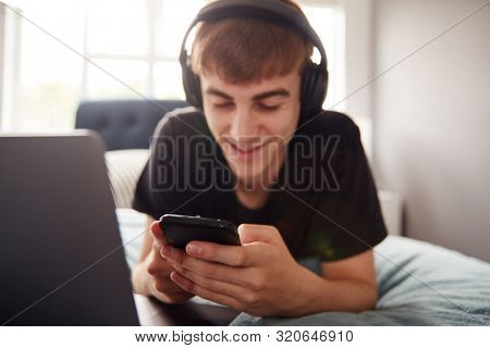 Male College Student Wearing Headphones Lies On Bed In Shared House With Mobile Phone Using Laptop