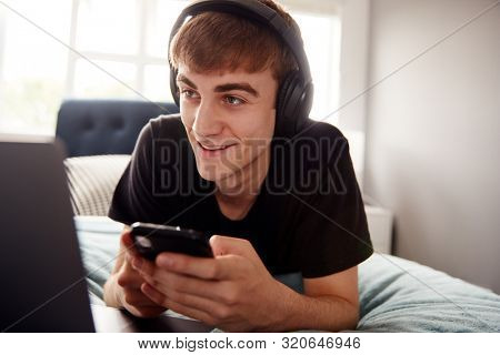 Male College Student Wearing Headphones Lies On Bed In Shared House With Mobile Phone Using Laptop