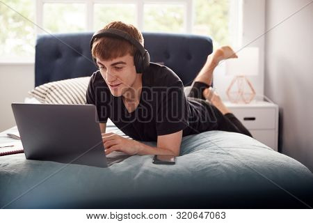 Male College Student Wearing Headphones Lies On Bed In Shared House Working On Laptop