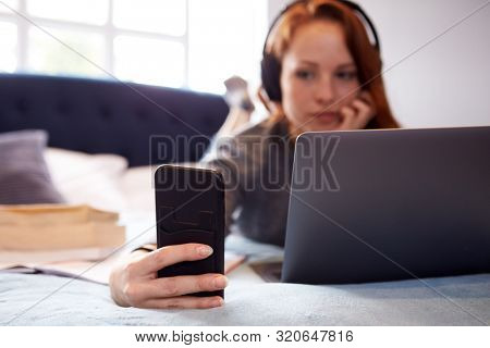 Female College Student Wearing Headphones Lies On Bed In Shared House With Mobile Phone Using Laptop