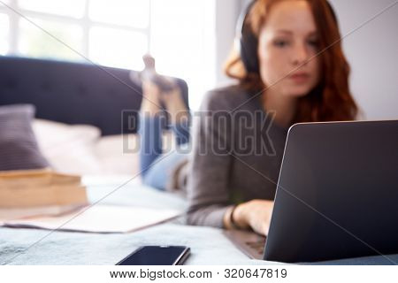 Female College Student Wearing Headphones Lies On Bed In Shared House With Mobile Phone Using Laptop