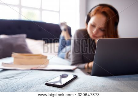 Female College Student Wearing Headphones Lies On Bed In Shared House With Mobile Phone Using Laptop