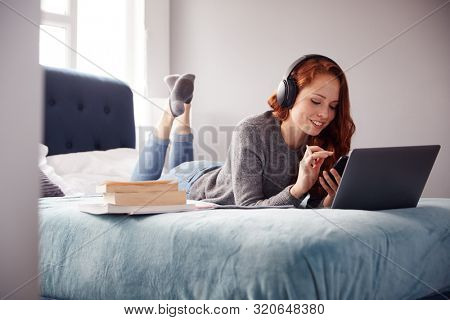 Female College Student Wearing Headphones Lies On Bed In Shared House With Mobile Phone Using Laptop