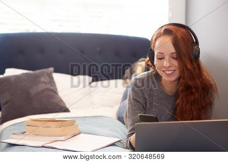 Female College Student Wearing Headphones Lies On Bed In Shared House With Mobile Phone Using Laptop
