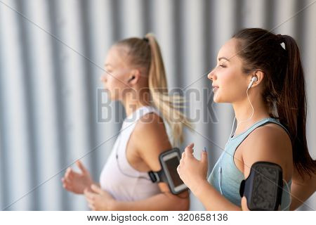 fitness, sport and healthy lifestyle concept - young women or female friends with earphones wearing armbands with smartphones and running outdoors
