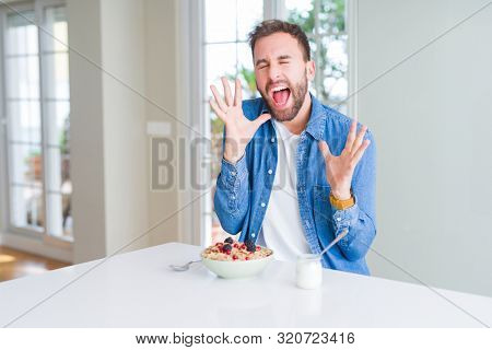 Handsome man eating cereals for breakfast at home celebrating mad and crazy for success with arms raised and closed eyes screaming excited. Winner concept