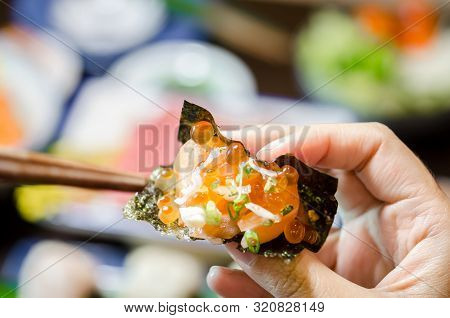 Hand Holding Salmon Sashimi With Sheet Of Dried Seaweed At Japanese Food Restaurant.