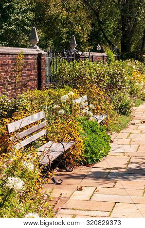 A Stone Walkway In A Garden Of Floers With Two Wooden Benches And A Brick Wall In Summer, Pittsburgh