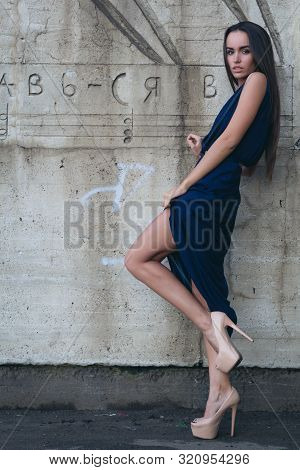 A Glamorous Woman In A Short Evening Dress With Heels Stands Next To The Concrete Wall.
